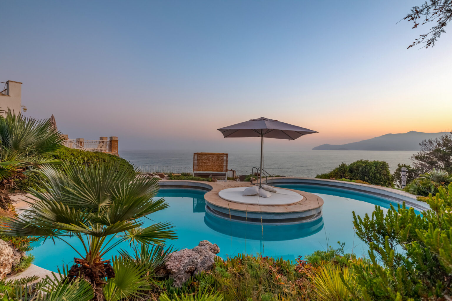 Scenic view of a luxury pool with a circular design, surrounded by lush greenery and a parasol, overlooking the ocean at sunset. Ideal for vacation rentals and luxury getaways. | Only on SopranoVillas