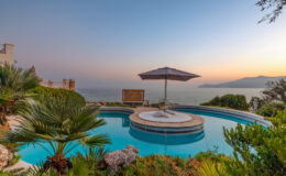 Scenic view of a luxury pool with a circular design, surrounded by lush greenery and a parasol, overlooking the ocean at sunset. Ideal for vacation rentals and luxury getaways.