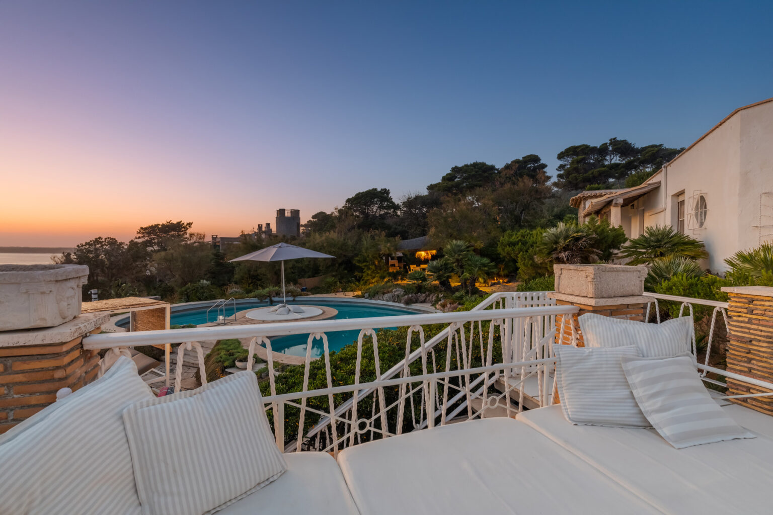 Luxurious villa with a stunning pool surrounded by lush gardens at sunset, featuring a cozy outdoor seating area with plush cushions. | Only on SopranoVillas