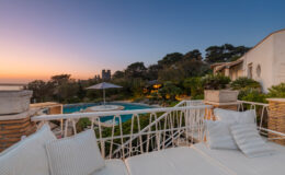 Luxurious villa with a stunning pool surrounded by lush gardens at sunset, featuring a cozy outdoor seating area with plush cushions.
