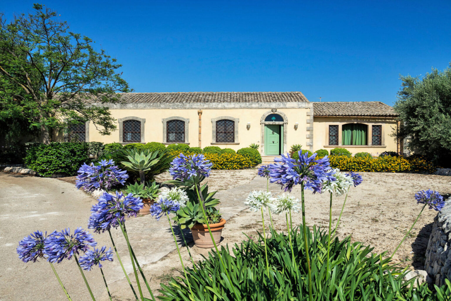 Charming countryside home with a stone facade, surrounded by vibrant flowers and lush greenery under a clear blue sky. | Only on SopranoVillas