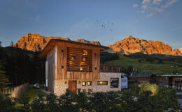 Modern wooden house surrounded by lush greenery and mountains at sunset, showcasing architectural design and natural beauty in a tranquil setting.