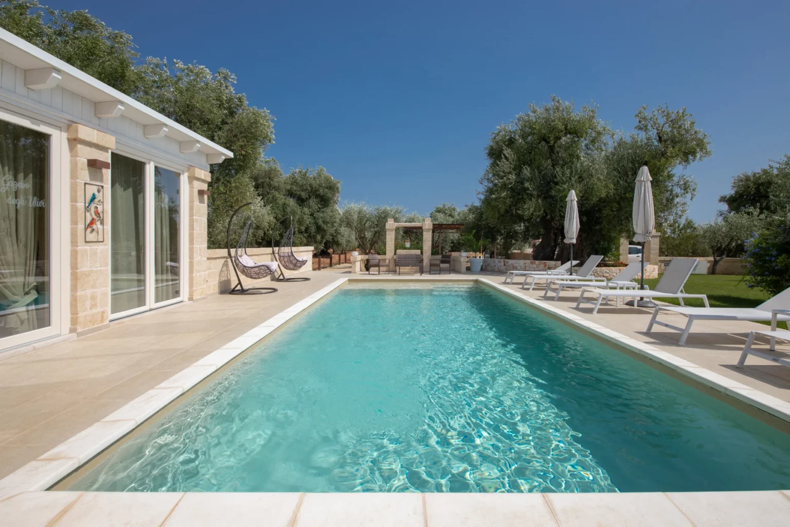 Luxurious outdoor pool area with modern lounge chairs, surrounded by olive trees and a clear blue sky, ideal for relaxation and summer gatherings. | Only on SopranoVillas
