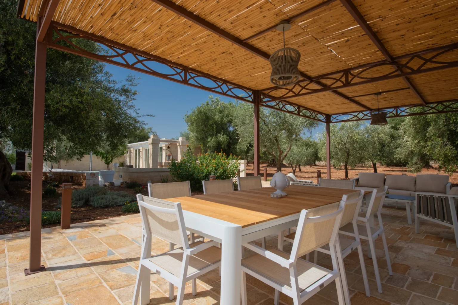 A modern outdoor dining area under a bamboo roof, featuring a wooden table surrounded by white chairs, with olive trees and landscaped gardens in the background. Ideal for alfresco dining and relaxation. | Only on SopranoVillas