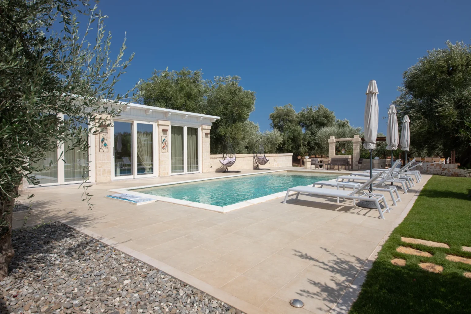 Modern villa with a swimming pool surrounded by olive trees, featuring lounge chairs and shaded areas for relaxation under a clear blue sky. Ideal for a tranquil getaway. | Only on SopranoVillas