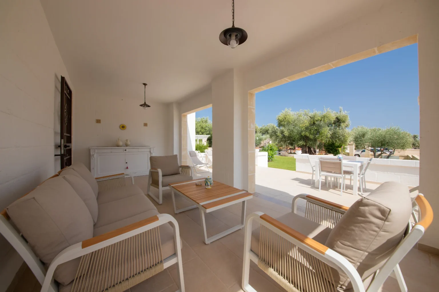Cozy outdoor living space featuring comfortable seating, a wooden coffee table, and a view of a lush garden with olive trees. The area is bright and airy, illuminated by natural light, creating a perfect spot for relaxation or entertaining. | Only on SopranoVillas