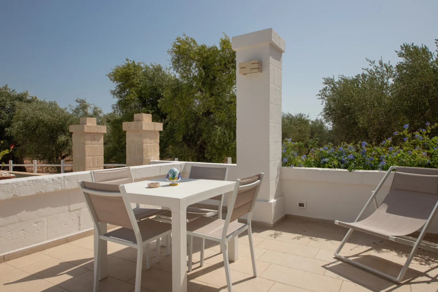 Spacious outdoor terrace featuring a white dining table with four chairs, two lounge chairs, and a scenic view of lush greenery and flowering plants, perfect for relaxation and dining al fresco. | Only on SopranoVillas