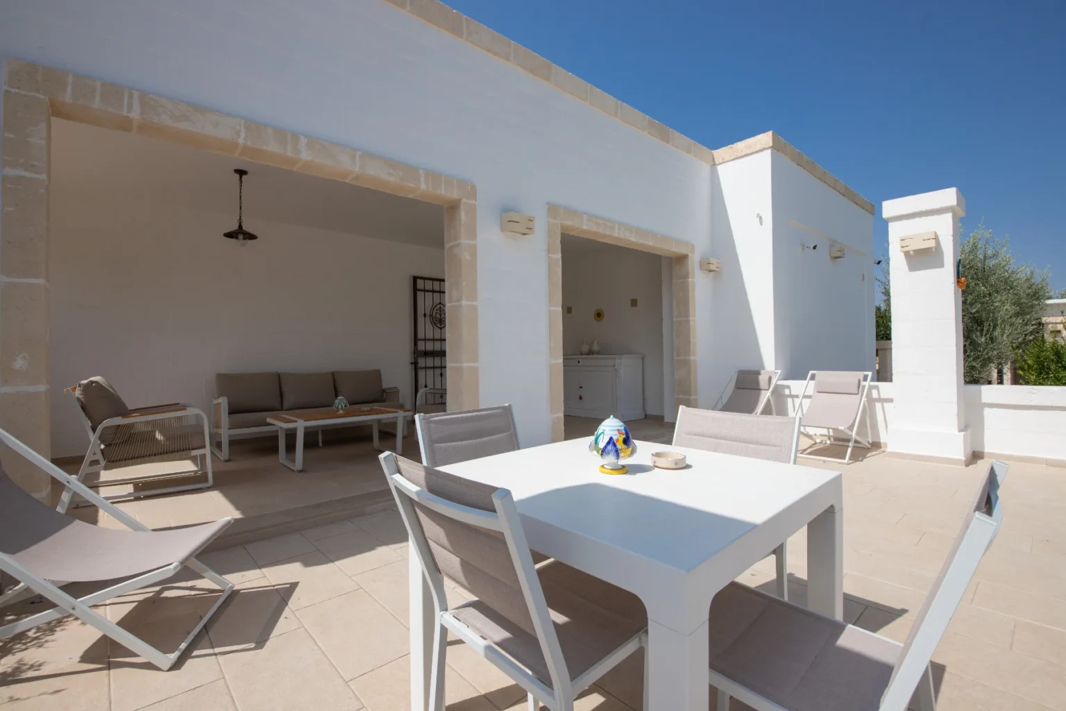 Modern outdoor patio with white walls and tiled flooring, featuring a dining table with chairs, lounge seating, and decorative accents, ideal for relaxation and entertaining in a sunny environment. | Only on SopranoVillas