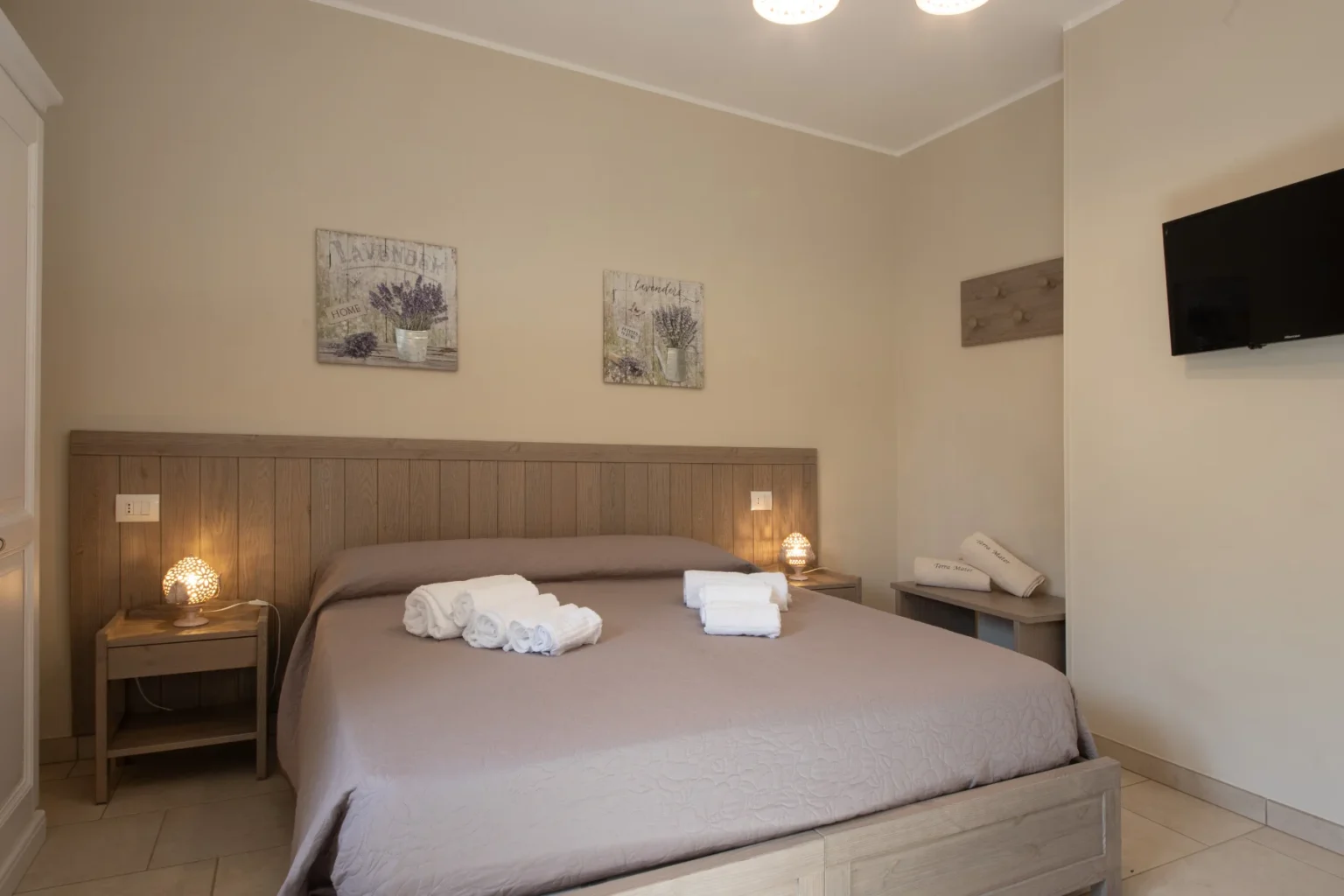 Cozy bedroom featuring a queen-sized bed with neutral bedding, decorative lavender artwork on the walls, a bedside lamp, and neatly arranged towels, creating a relaxing atmosphere. The room includes a wall-mounted TV and a minimalist design with wooden accents. | Only on SopranoVillas