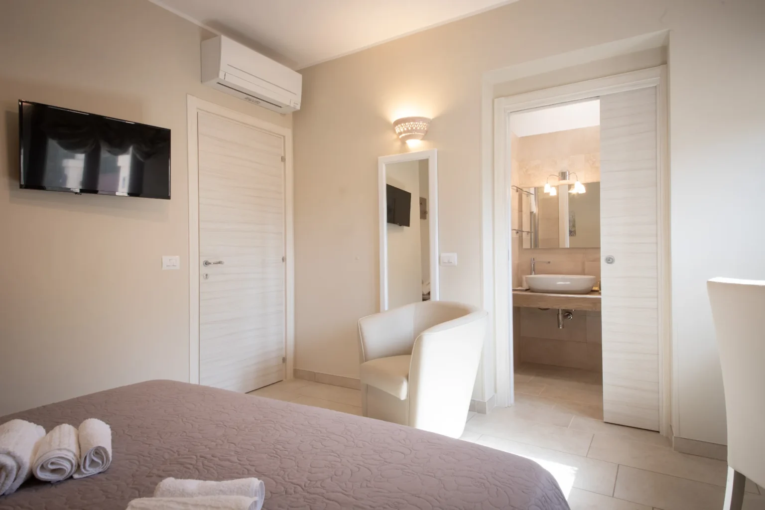 Cozy hotel room featuring a comfortable bed with rolled towels, a wall-mounted TV, and an en-suite bathroom. Modern decor includes a stylish armchair and neutral tones, creating a welcoming atmosphere for guests. | Only on SopranoVillas