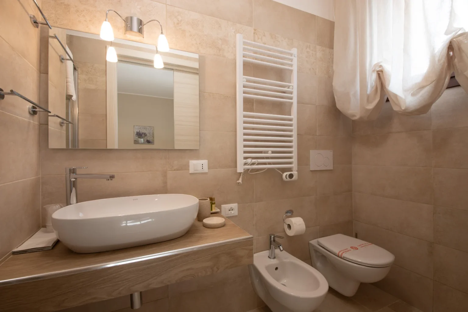 Modern bathroom featuring a sleek oval sink, wooden countertop, and stylish fixtures. The space includes a bidet, toilet, and a towel warmer, with natural light filtering through sheer curtains. Neutral-toned tiles enhance the contemporary design. | Only on SopranoVillas