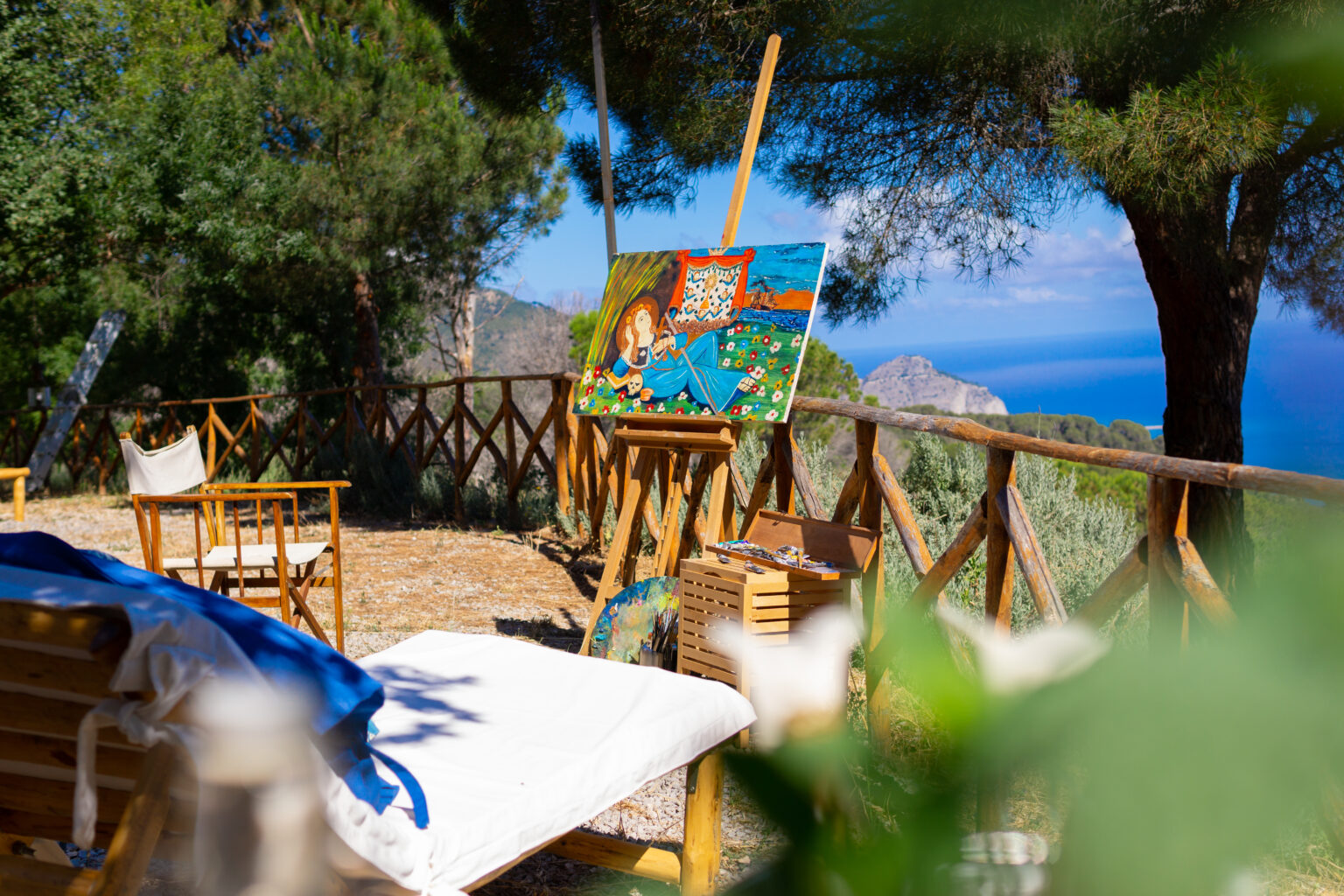Scenic outdoor art studio with an easel displaying a colorful painting, surrounded by wooden chairs and lush greenery, overlooking a tranquil sea view. | Only on SopranoVillas