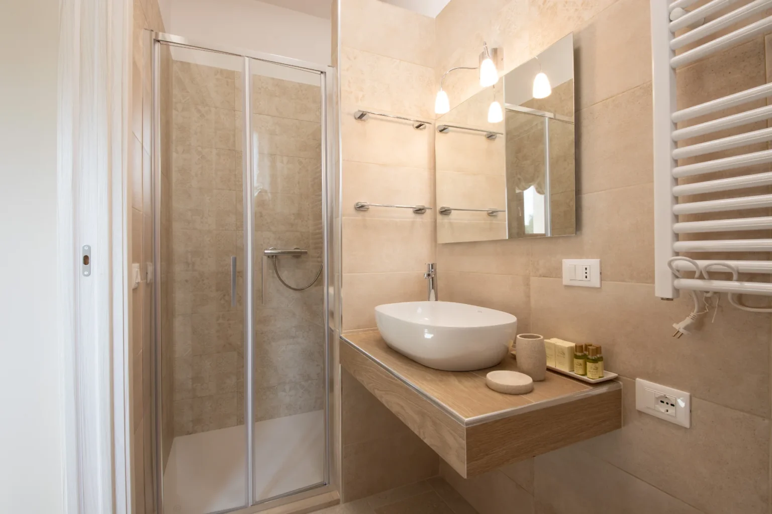 Modern bathroom interior featuring a glass shower enclosure, a stylish oval sink on a wooden countertop, and beige tiled walls. The space is illuminated by contemporary light fixtures and includes neatly arranged toiletries and a heated towel rack. | Only on SopranoVillas