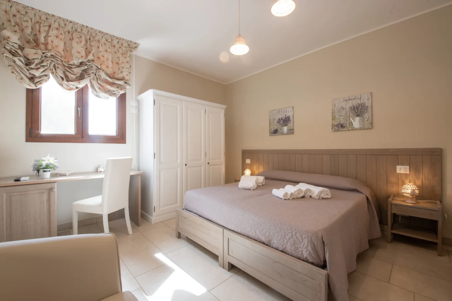 Cozy bedroom interior featuring a queen-sized bed with folded towels, a white wardrobe, and a wooden desk with a chair. Natural light streams through a window adorned with floral curtains, creating a warm and inviting atmosphere. Ideal for relaxation or work. | Only on SopranoVillas