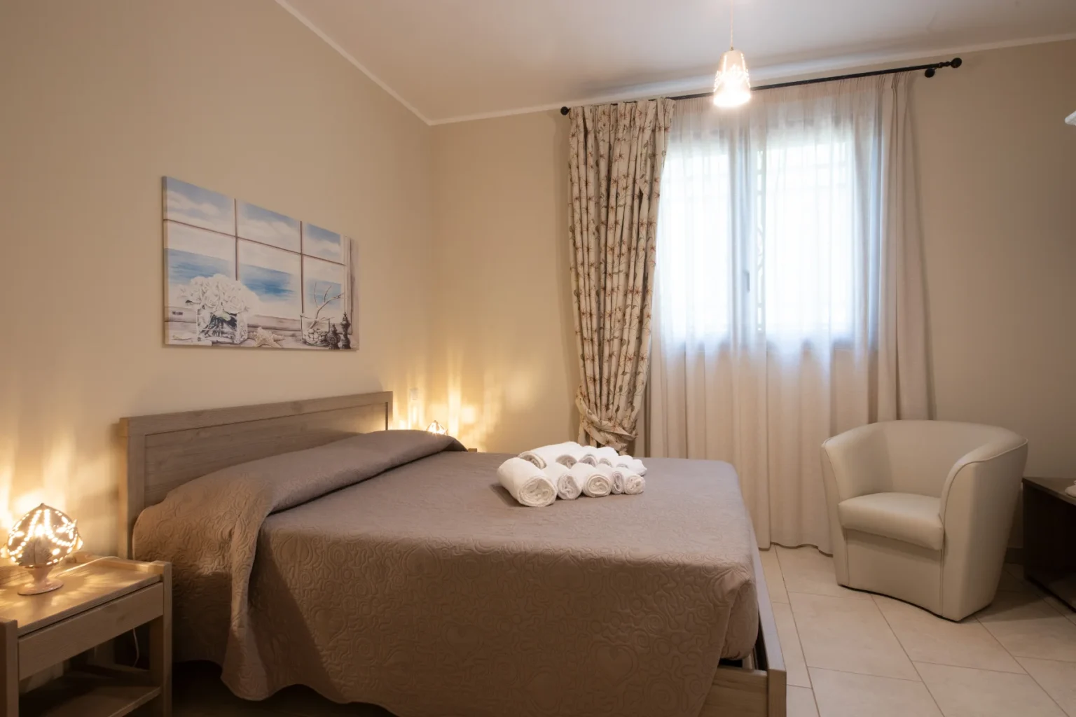 Cozy bedroom featuring a comfortable queen-sized bed with a decorative quilt, neatly arranged towels, a stylish armchair, and soft ambient lighting. The room is adorned with a scenic wall art piece and sheer curtains allowing natural light to fill the space, creating a warm and inviting atmosphere. | Only on SopranoVillas