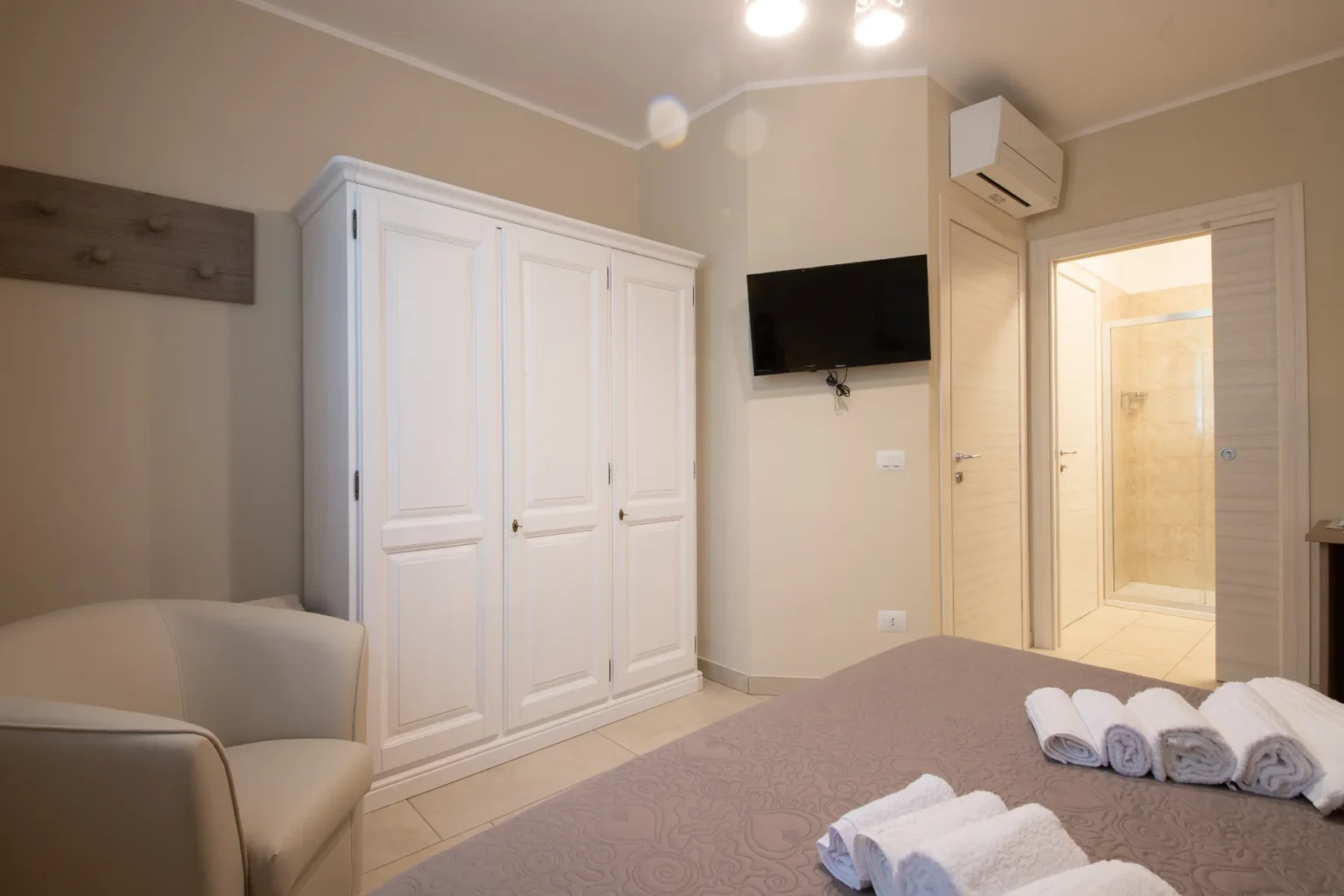 Modern bedroom interior featuring a comfortable armchair, a spacious white wardrobe, a wall-mounted TV, and neatly arranged towels on the bed, with an ensuite bathroom visible in the background. | Only on SopranoVillas