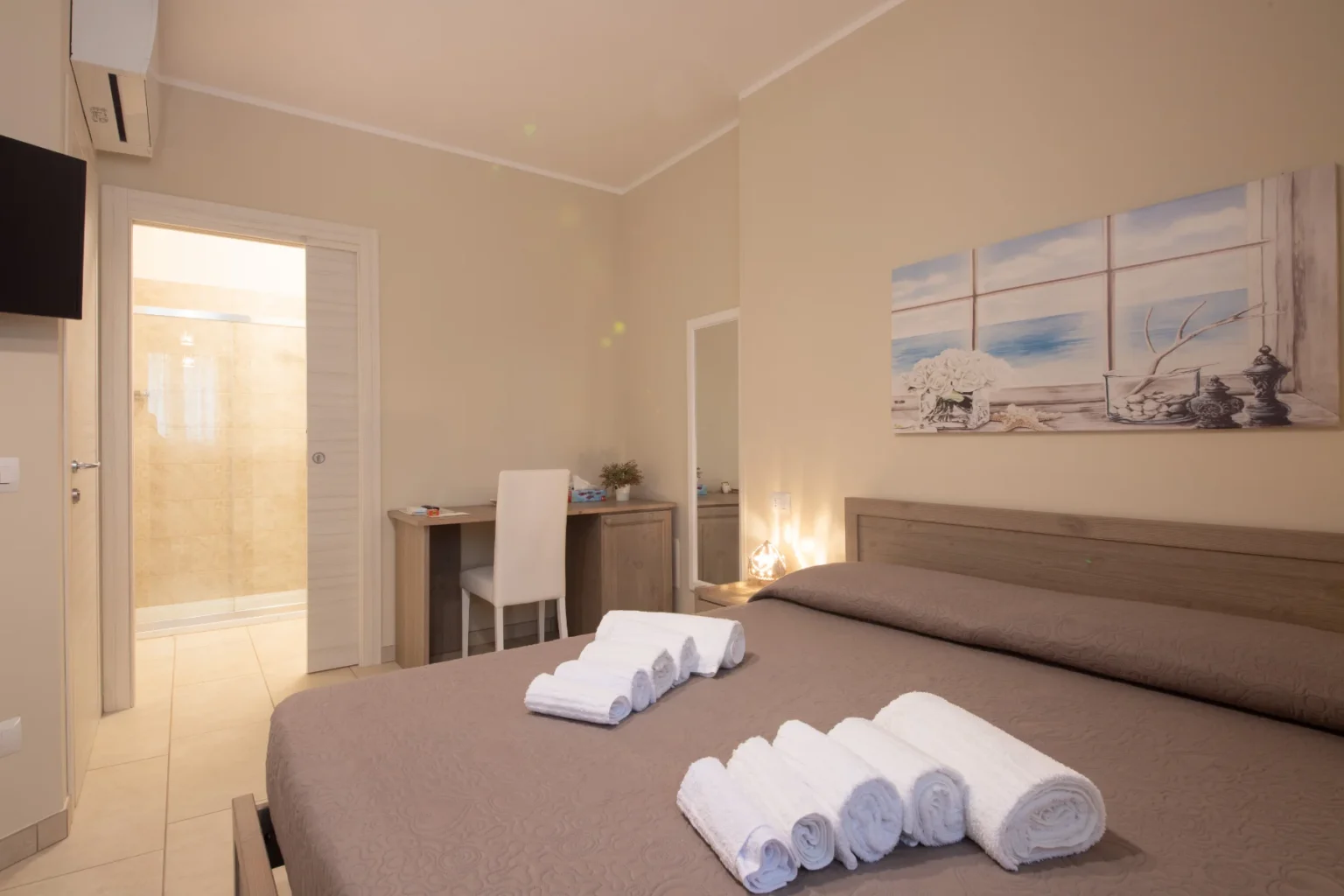 Cozy hotel room featuring a large bed with neatly arranged towels, a desk with a chair, and a bathroom with a glass shower. The room has a warm color palette and is decorated with a beach-themed artwork, creating a relaxing atmosphere. | Only on SopranoVillas