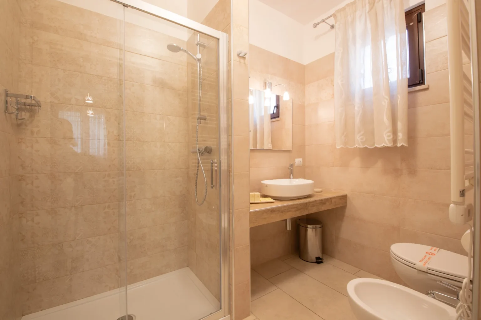 Modern bathroom featuring a glass shower enclosure, a round sink on a wooden countertop, and neutral-toned tile walls. The space includes a window with sheer curtains, a waste bin, and a toilet, creating a clean and inviting atmosphere. | Only on SopranoVillas