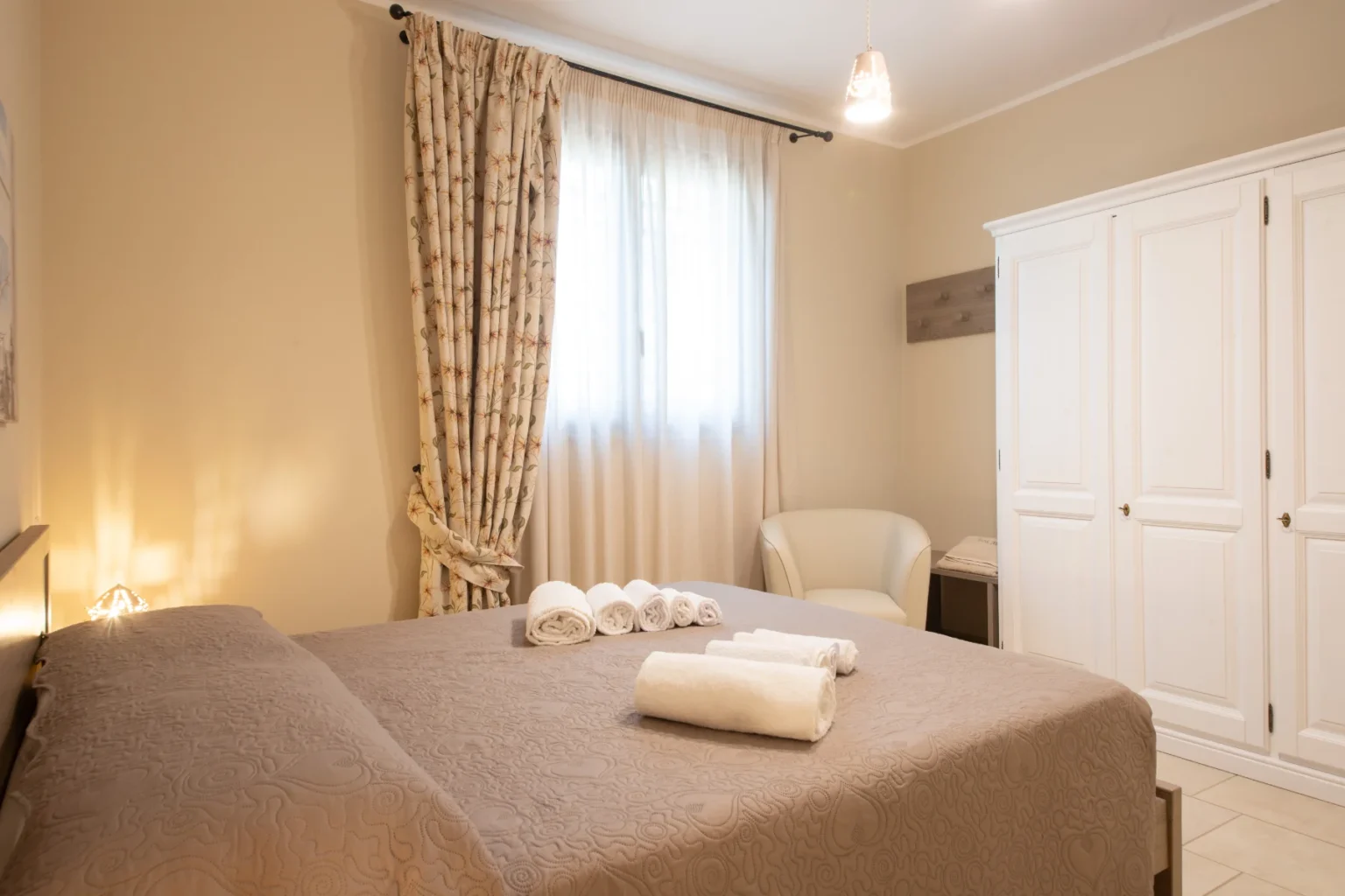 Cozy bedroom featuring a large bed with decorative bedding, neatly arranged white towels, a comfortable armchair, and a wardrobe. Natural light filters through sheer curtains, creating a warm and inviting atmosphere. | Only on SopranoVillas