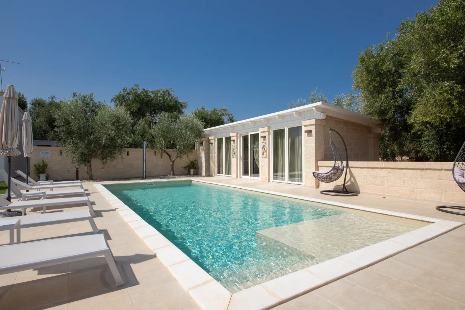 Modern villa with a private swimming pool surrounded by olive trees, featuring sun loungers and stylish hanging chairs, set against a clear blue sky. Ideal for relaxation and outdoor living. | Only on SopranoVillas