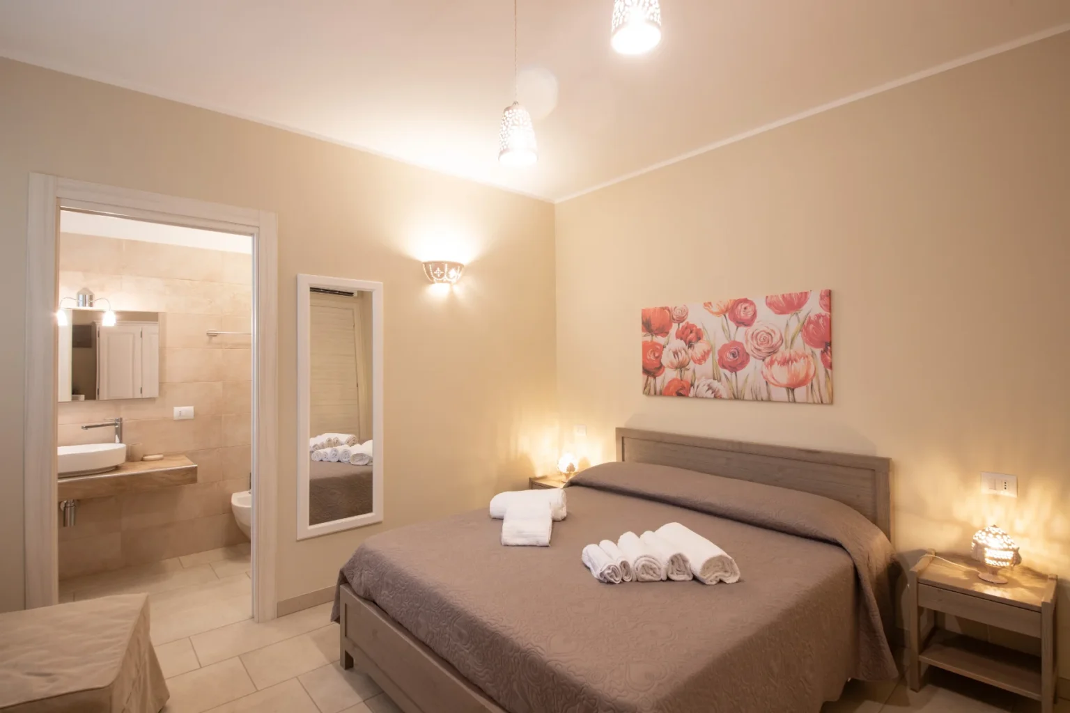 Cozy bedroom featuring a queen-size bed with neatly arranged towels, soft lighting, and a floral wall art piece. The room includes a mirror and an open doorway leading to a modern bathroom with elegant fixtures. Ideal for a relaxing stay. | Only on SopranoVillas