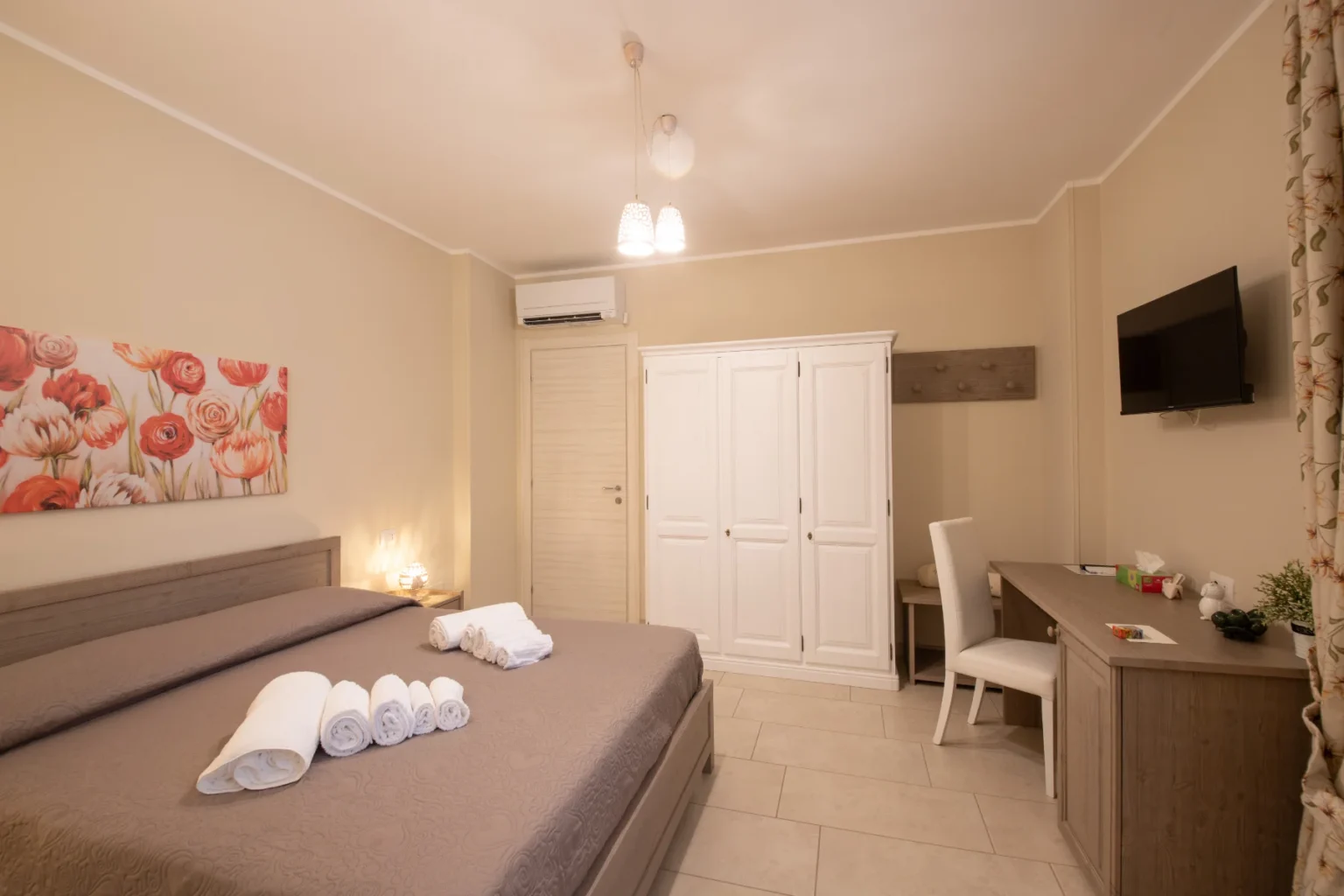 Cozy hotel room featuring a large bed with decorative pillows, neatly arranged towels, a wooden desk and chair, a flat-screen TV, and a wardrobe, all complemented by soft lighting and floral artwork on the wall. | Only on SopranoVillas