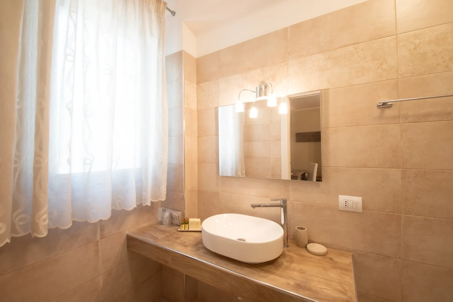 Modern bathroom interior featuring a stylish white vessel sink on a wooden vanity, beige tiled walls, and a large mirror illuminated by contemporary light fixtures. Natural light filters through sheer curtains, creating a bright and inviting atmosphere. | Only on SopranoVillas