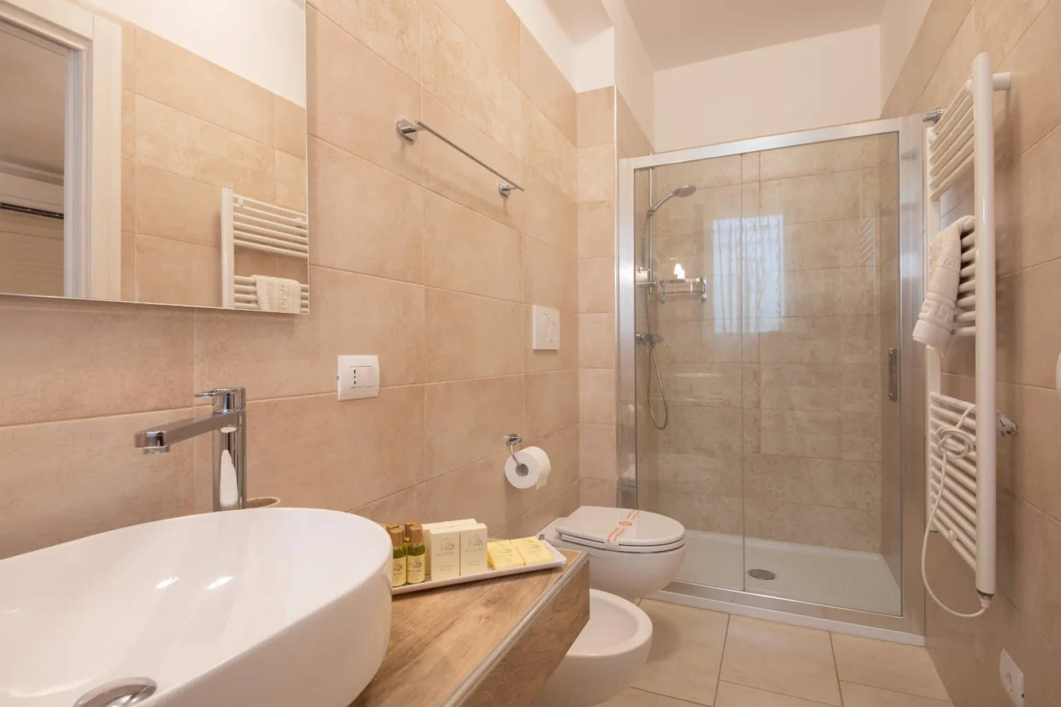 Modern bathroom featuring a walk-in shower, sleek white sink, wooden vanity, and complimentary toiletries. Neutral-toned tiles enhance the spacious and clean aesthetic. | Only on SopranoVillas