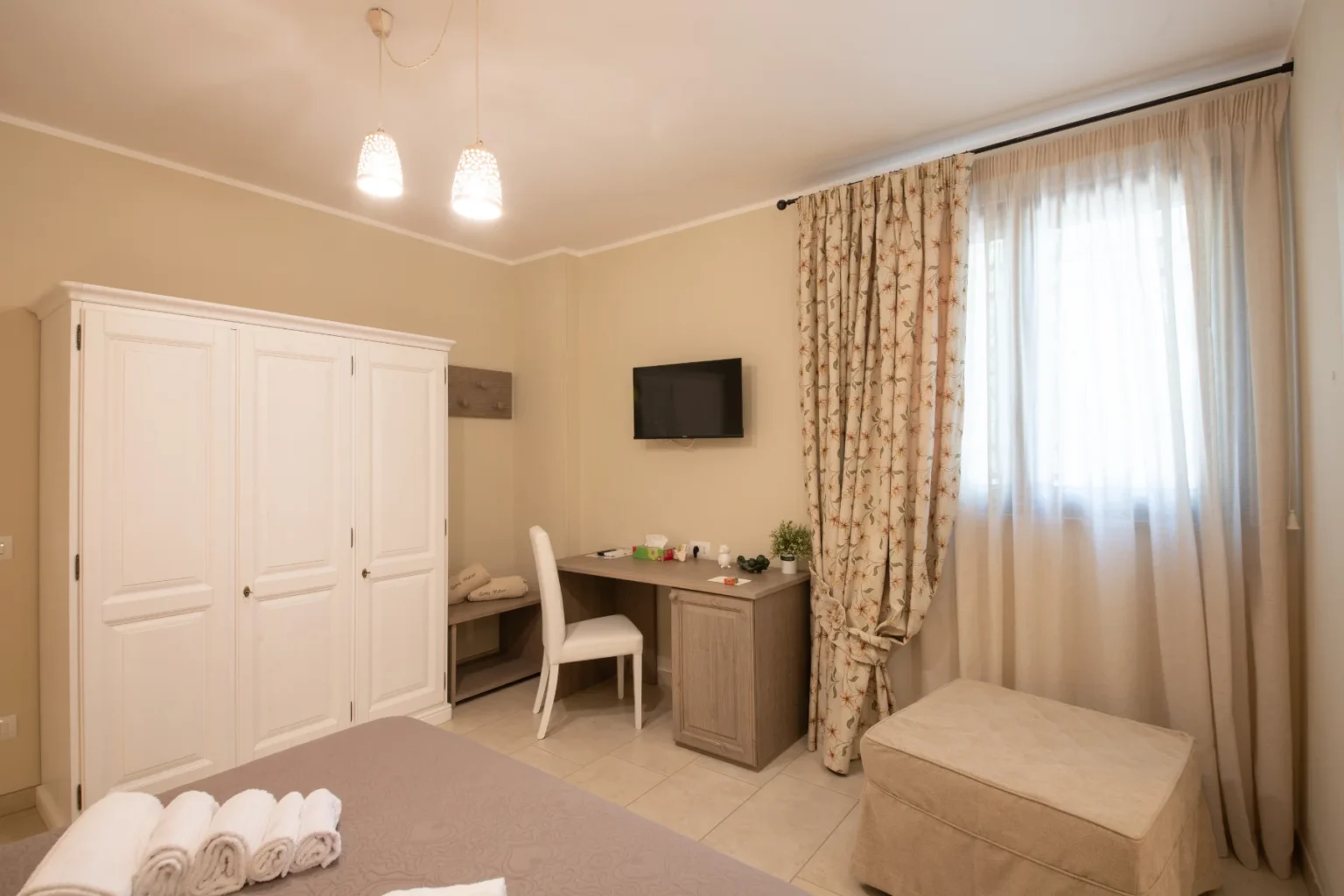 Cozy hotel room featuring a comfortable bed with neatly arranged towels, a stylish desk with a chair, a flat-screen TV mounted on the wall, and elegant curtains. The room is illuminated by modern pendant lights and has a spacious wardrobe, creating a warm and inviting atmosphere. | Only on SopranoVillas