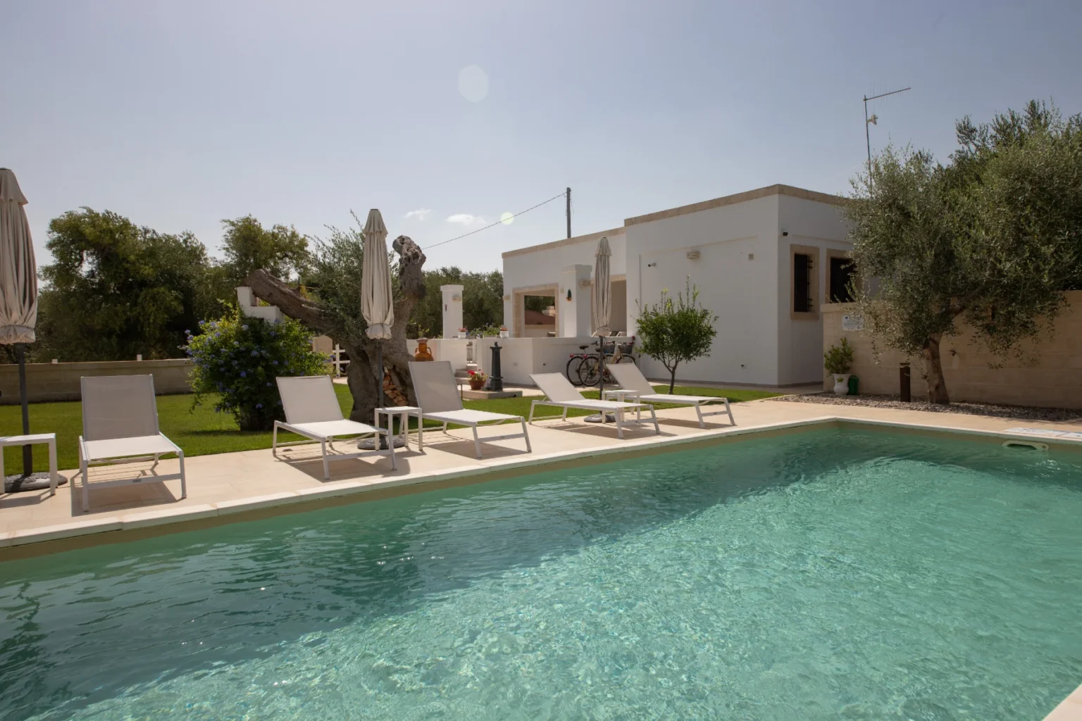 Modern villa with a swimming pool surrounded by sun loungers and umbrellas, set in a lush garden with olive trees under a clear blue sky. Ideal for relaxation and outdoor gatherings. | Only on SopranoVillas