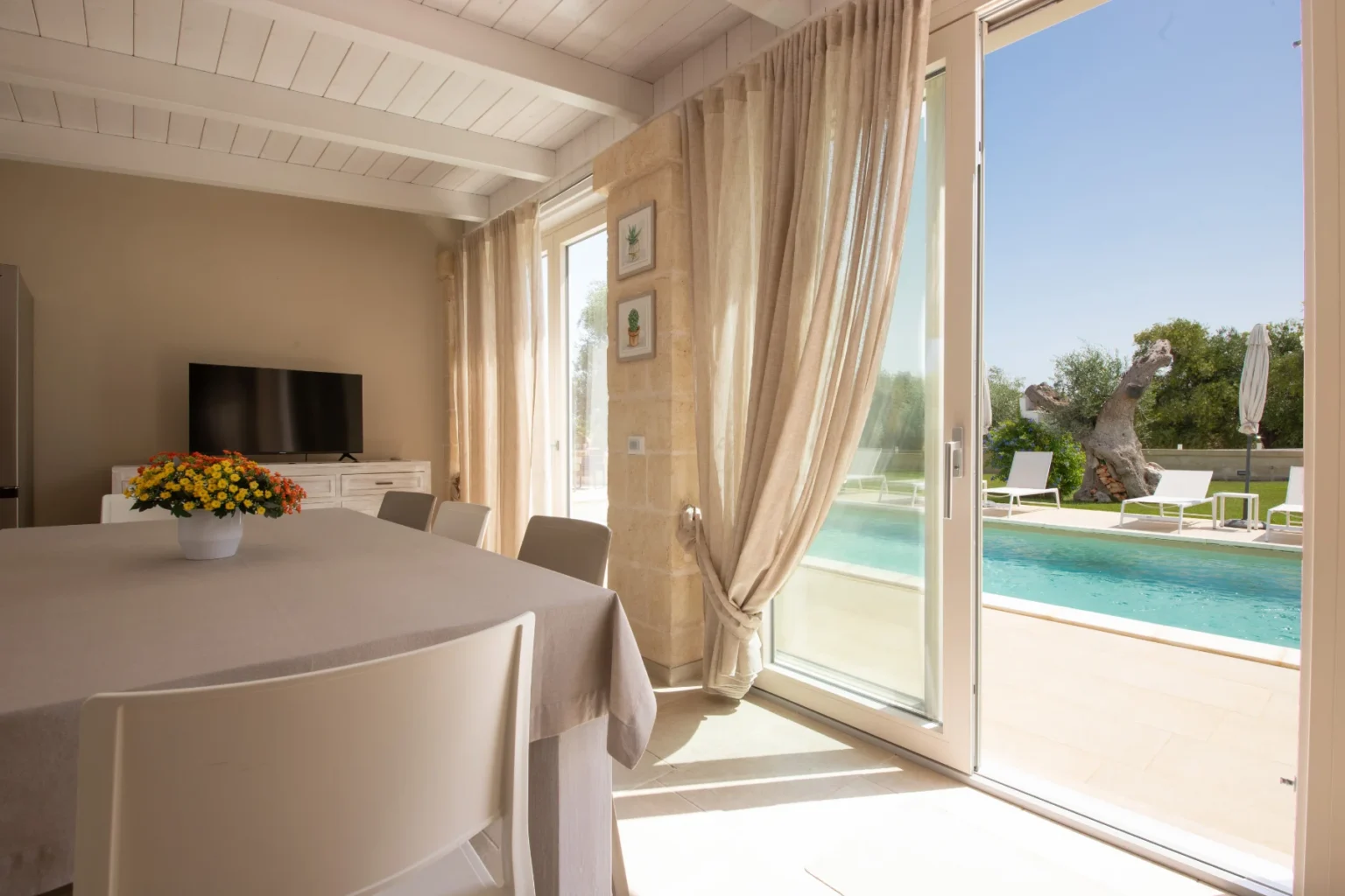 Bright and airy dining area featuring a light-colored table, white chairs, and a vibrant flower centerpiece, with large glass doors leading to a poolside view and outdoor seating. Natural light floods the space, highlighting the modern decor and serene ambiance. | Only on SopranoVillas