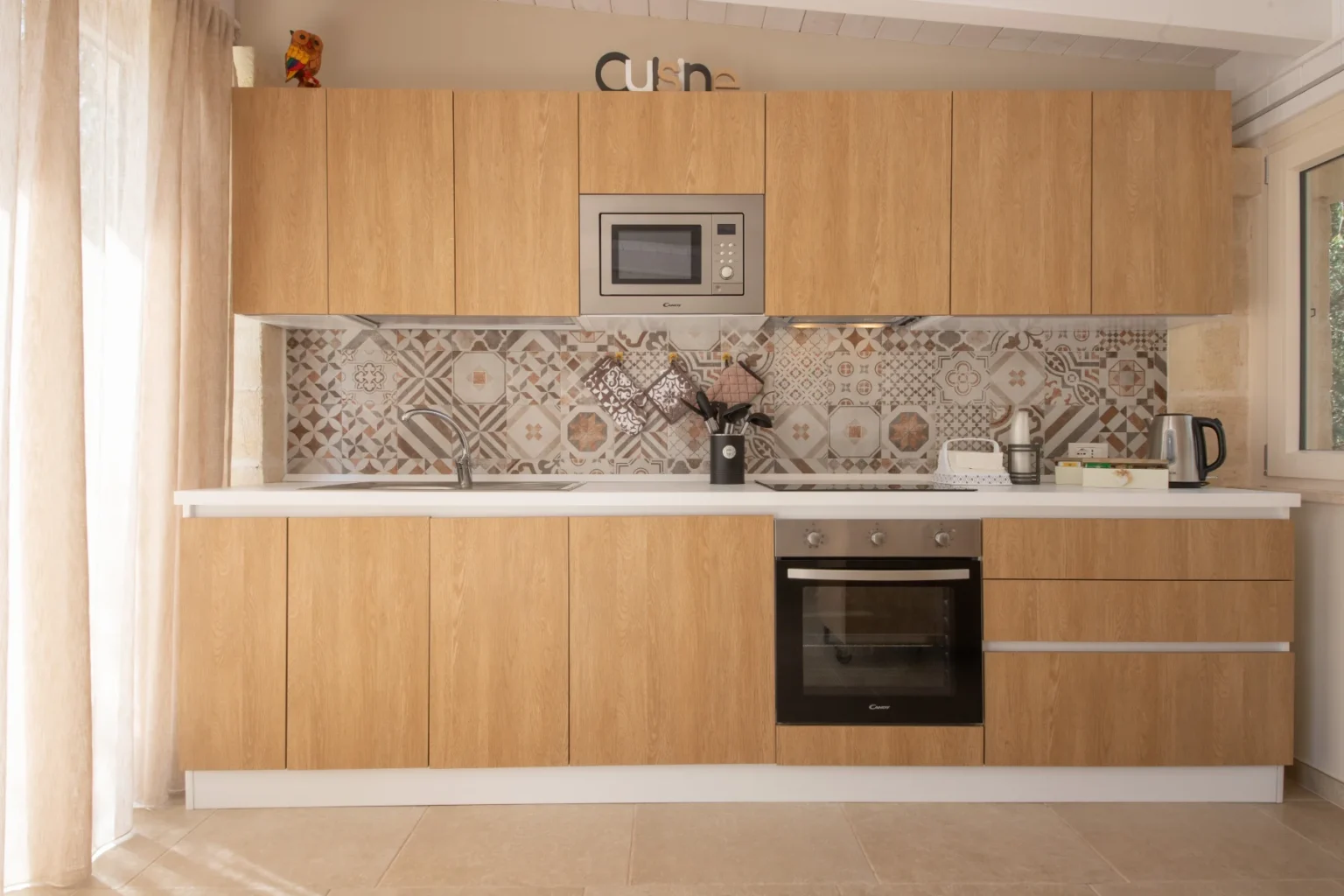 Modern kitchen design featuring wooden cabinetry, a microwave, and a patterned backsplash. The space includes a sink, stovetop, and oven, complemented by a kettle and cooking utensils. Natural light filters through sheer curtains, creating a warm and inviting atmosphere. | Only on SopranoVillas