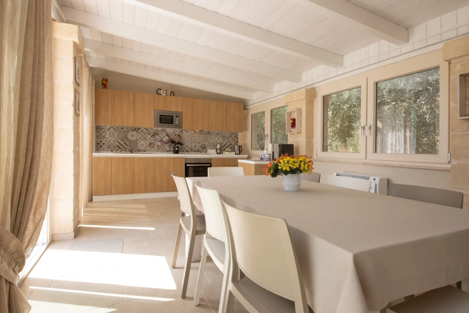 Bright and airy dining area featuring a large table with a neutral tablecloth, surrounded by white chairs. The modern kitchen showcases wooden cabinetry, a patterned backsplash, and ample natural light streaming through large windows, creating a warm and inviting atmosphere. | Only on SopranoVillas