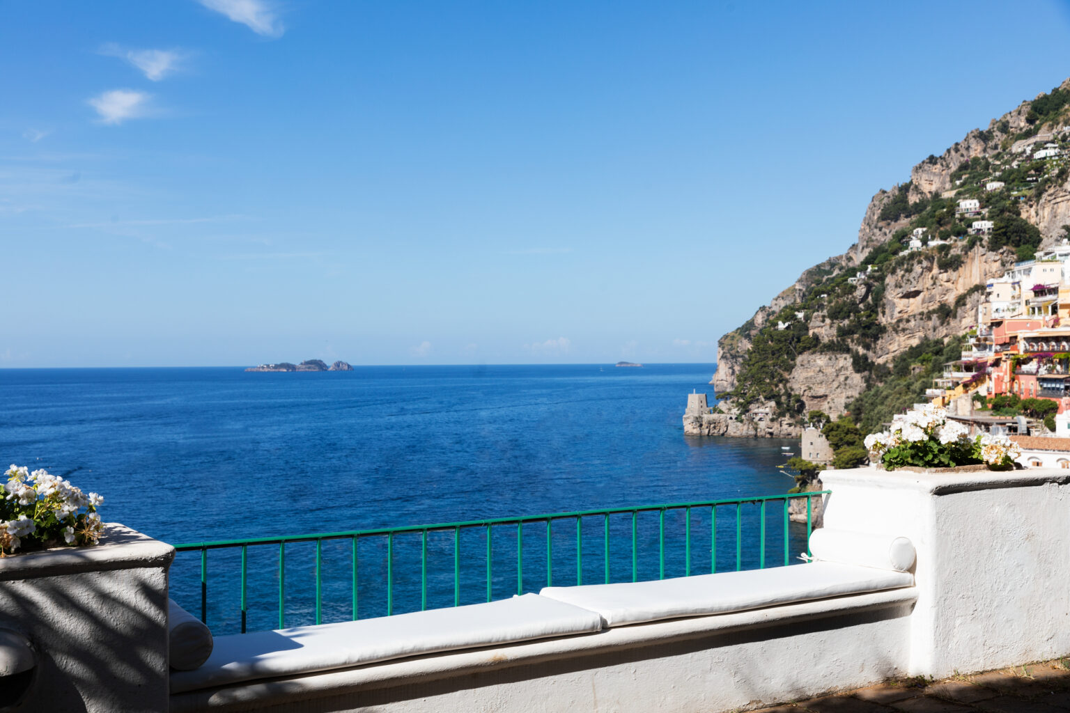 Scenic view of the Amalfi Coast featuring a clear blue sea, rocky cliffs, and a vibrant sky, with a balcony railing in the foreground adorned with flowers. | Only on SopranoVillas
