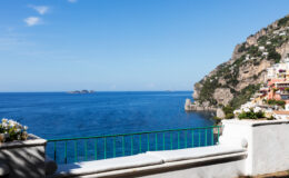 Scenic view of the Amalfi Coast featuring a clear blue sea, rocky cliffs, and a vibrant sky, with a balcony railing in the foreground adorned with flowers.