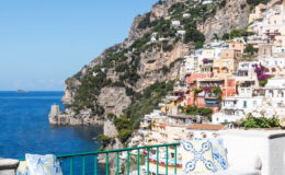 A picturesque view of Positano, Italy, showcasing colorful cliffside houses against a backdrop of clear blue skies and the Mediterranean Sea. The image features lush greenery on the hillside and traditional Italian architecture, highlighting the coastal charm of the Amalfi Coast.