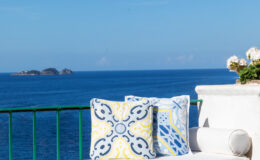 Colorful decorative pillows on a white terrace overlooking a serene blue ocean and distant islands, showcasing a tranquil coastal getaway.