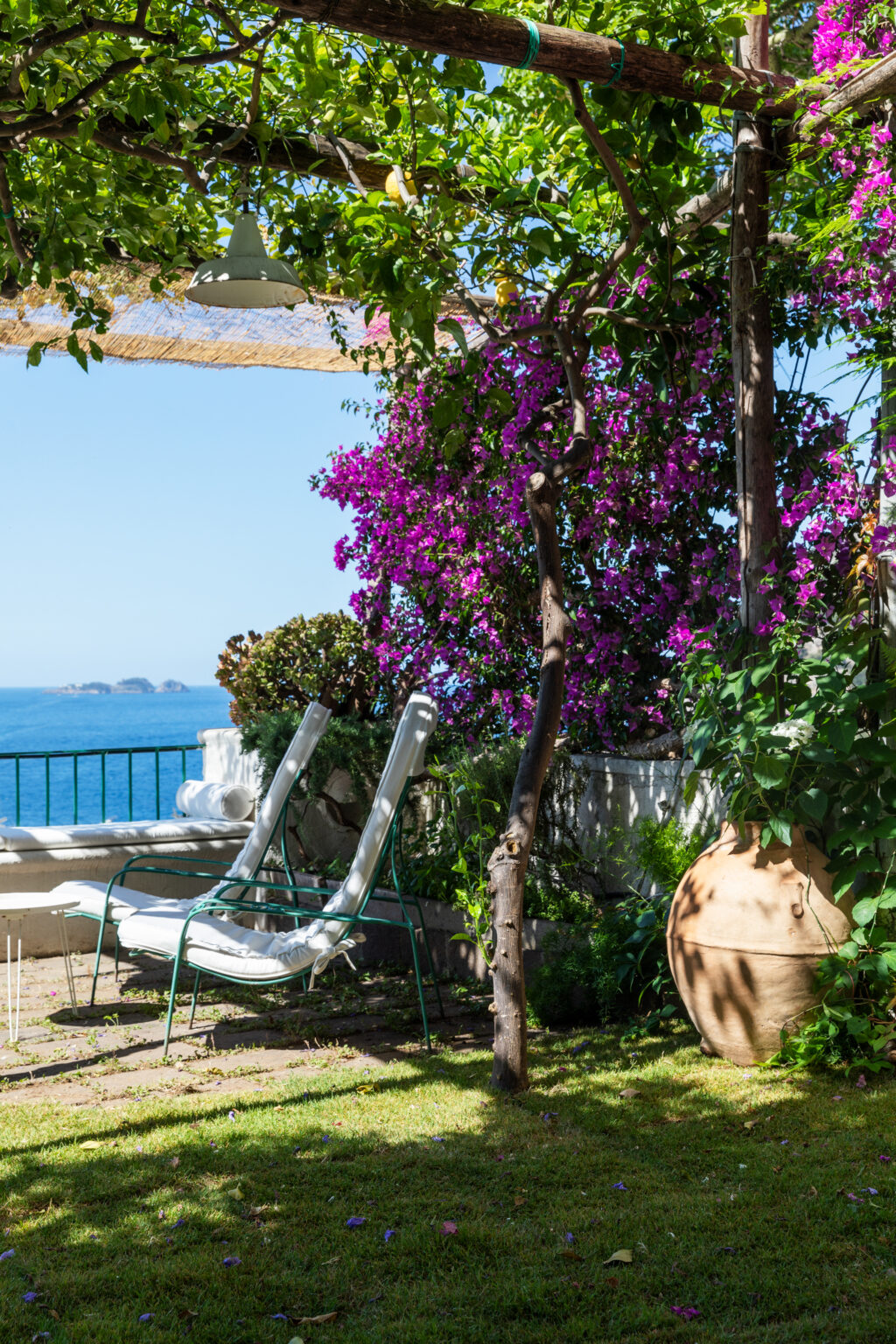 A serene outdoor garden with two white lounge chairs under a wooden pergola, surrounded by vibrant purple bougainvillea and lush greenery, overlooking a tranquil blue sea. | Only on SopranoVillas