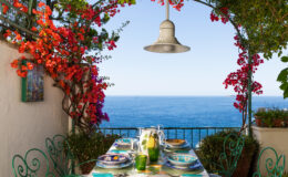 A picturesque outdoor dining area featuring a table set for a meal, surrounded by vibrant bougainvillea flowers and overlooking a serene ocean view. The setting includes colorful plates and glasses, with a pendant light hanging above, creating a relaxing and inviting atmosphere.
