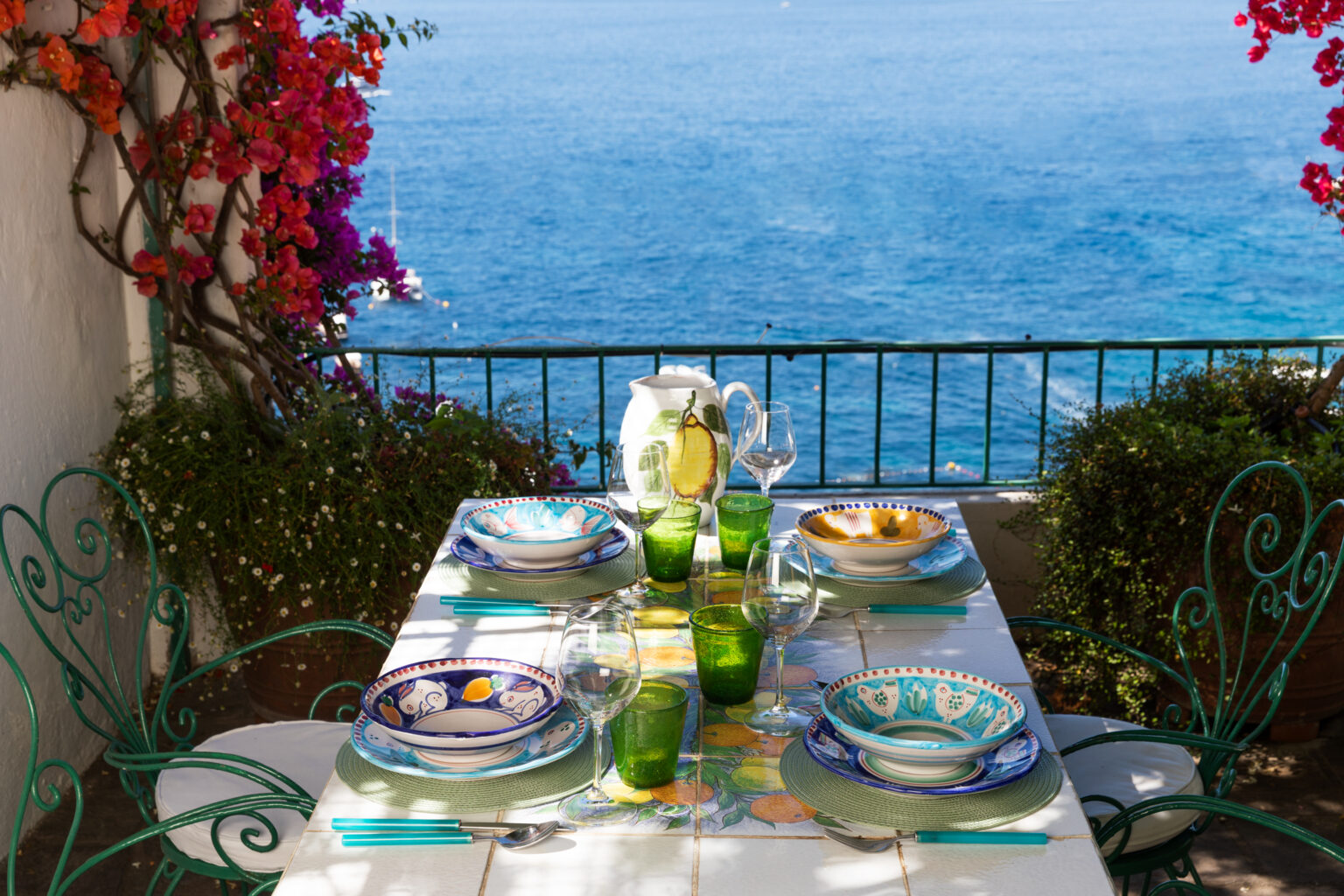 A beautifully set outdoor dining table with colorful plates, green glasses, and a pitcher, overlooking a serene blue ocean. Bougainvillea blooms add vibrant color to the scene, creating a picturesque dining experience. | Only on SopranoVillas