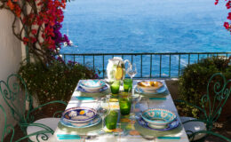 A beautifully set outdoor dining table with colorful plates, green glasses, and a pitcher, overlooking a serene blue ocean. Bougainvillea blooms add vibrant color to the scene, creating a picturesque dining experience.