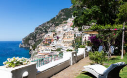 Scenic view of Positano, Italy, showcasing colorful cliffside houses, lush greenery, and a tranquil blue sea under a clear sky. The image captures a garden terrace with white flowers and lounge chairs, emphasizing the beauty of the Amalfi Coast.