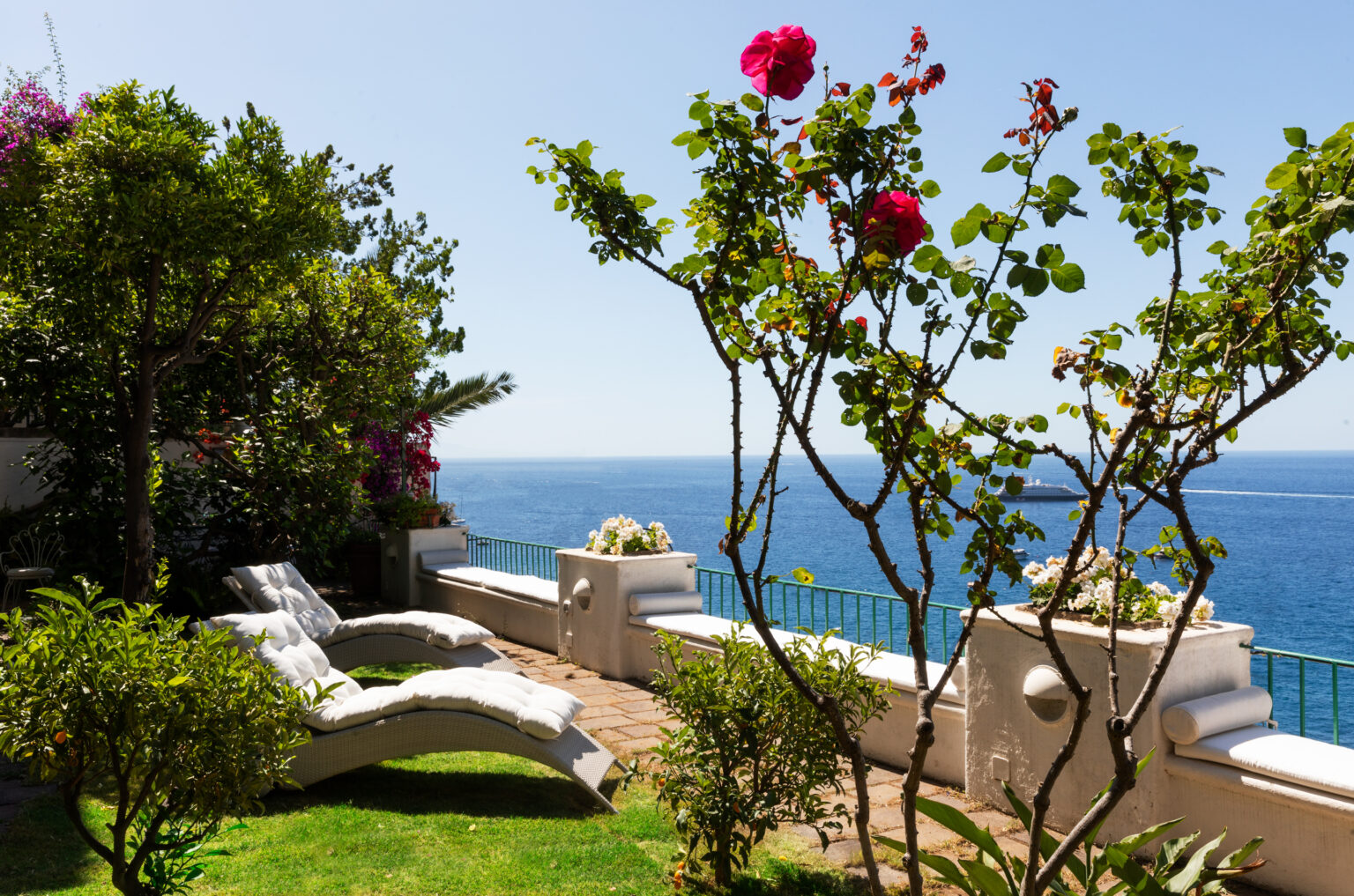 Lush garden with vibrant flowers, two lounge chairs, and a stunning ocean view under a clear blue sky, perfect for relaxation and outdoor enjoyment. | Only on SopranoVillas