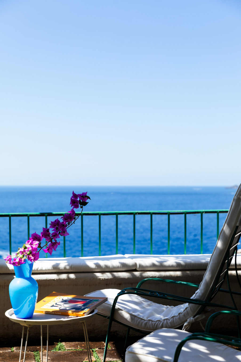 Vibrant purple flowers in a pot on a balcony overlooking a serene blue ocean under a clear sky. | Only on SopranoVillas