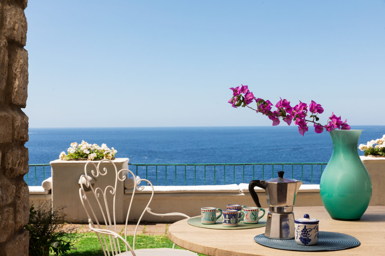Scenic ocean view from a terrace featuring a coffee setup with a moka pot, decorative cups, and a vibrant turquoise vase with blooming purple flowers, creating a serene and inviting atmosphere. | Only on SopranoVillas