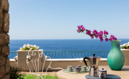 Scenic ocean view from a terrace featuring a coffee setup with a moka pot, decorative cups, and a vibrant turquoise vase with blooming purple flowers, creating a serene and inviting atmosphere.