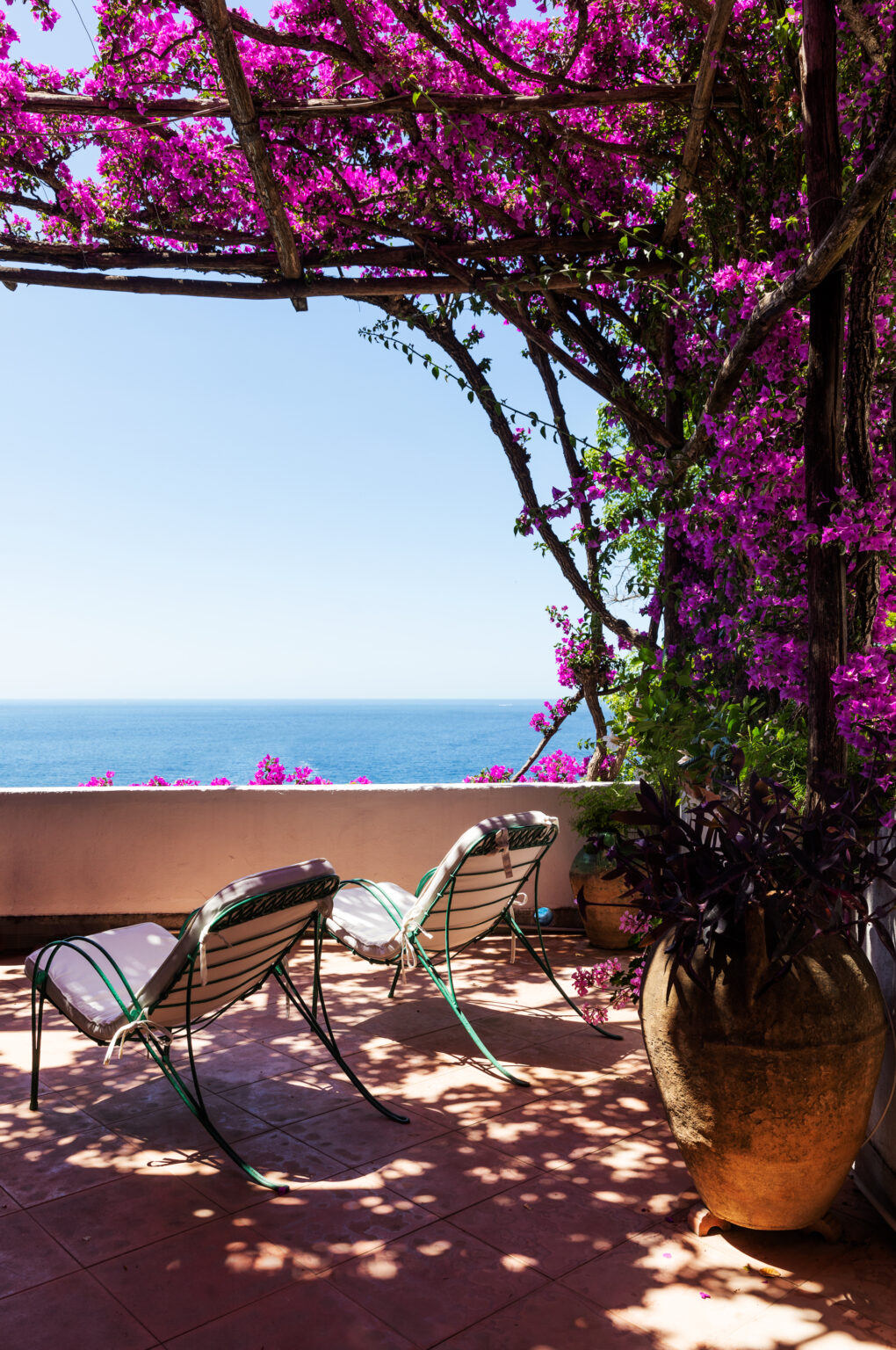 Cozy outdoor balcony with two lounge chairs surrounded by vibrant purple bougainvillea flowers, overlooking a serene ocean view under a clear blue sky. | Only on SopranoVillas