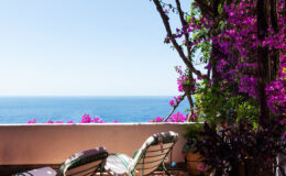 Cozy outdoor balcony with two lounge chairs surrounded by vibrant purple bougainvillea flowers, overlooking a serene ocean view under a clear blue sky.