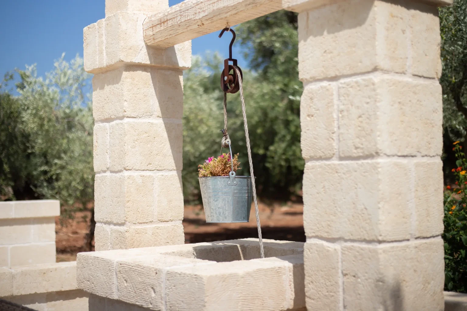 Rustic stone well with a metal bucket hanging by a pulley, adorned with colorful flowers, set against a backdrop of olive trees and clear blue sky. | Only on SopranoVillas
