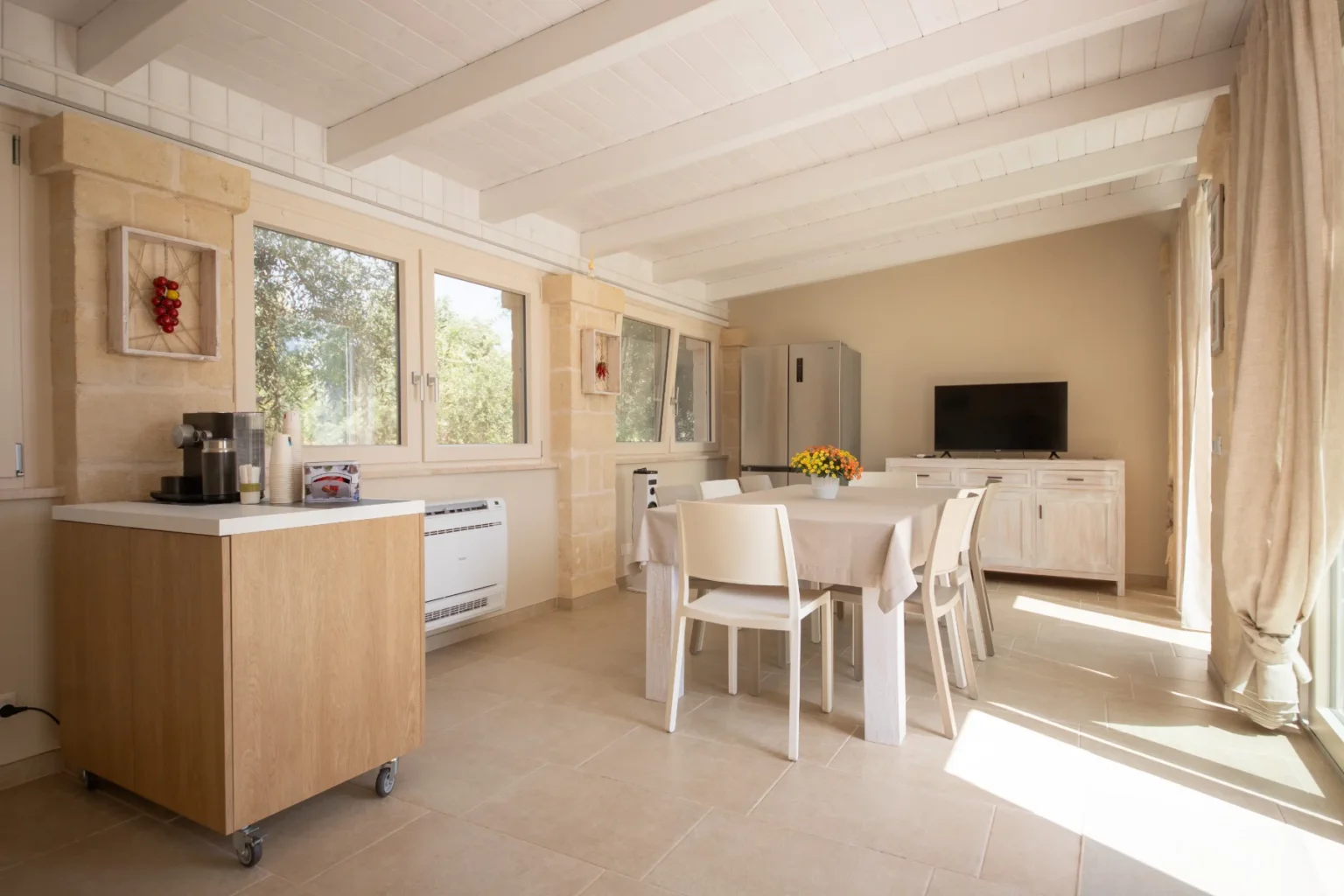 Bright and airy kitchen interior featuring a dining table, white chairs, and a modern refrigerator. The space includes a wooden coffee station and large windows allowing natural light, with decorative elements like a flower vase and wall art. | Only on SopranoVillas